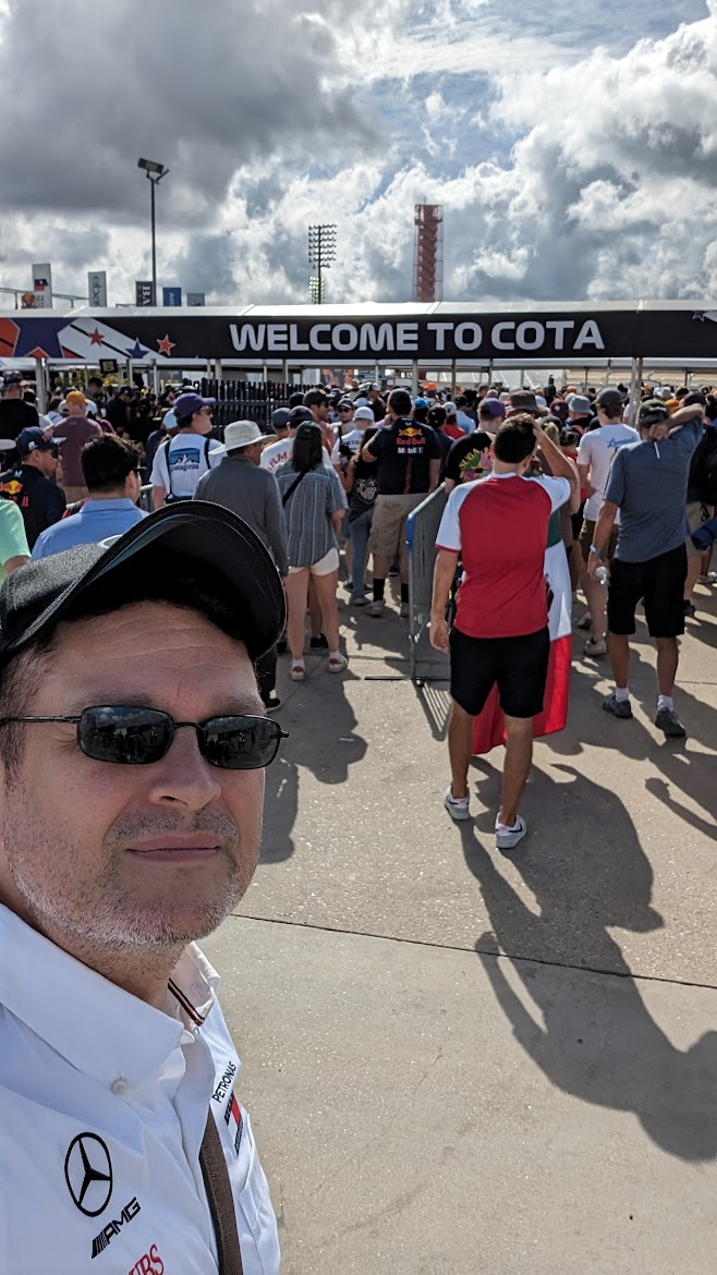 The photo shows a lively scene at the entrance of the **Circuit of the Americas (COTA)** racetrack in Austin, Texas.
In the foreground, a man is taking a selfie. He’s wearing a **white Mercedes-AMG team shirt**, a **black baseball cap**, and **glasses**. Behind him, there’s a **large crowd** of people waiting to enter the venue. Above the entrance, a big sign reads **“WELCOME TO COTA.”**
The atmosphere is energetic and sunny — many fans are dressed in **team shirts, caps, and sunglasses**, representing various Formula 1 teams like Red Bull and Mercedes. The **sky is partly cloudy**, with bright sunlight breaking through, creating a warm and vivid feel typical of a race day.
In the background, you can see **tall floodlights** and the **red observation tower**, a distinctive landmark of the COTA racetrack.