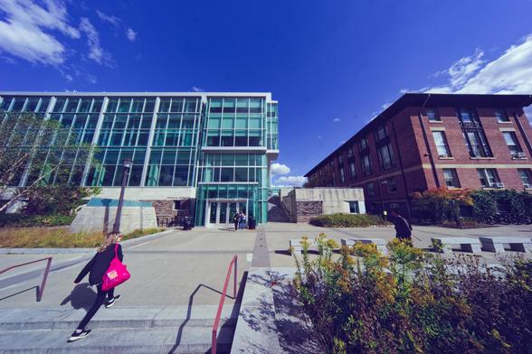 Slightly more than half of the image on the right is a short-looking glass and steel building, and on the right a brick building of about the same height with some small concrete stairs structure between them,  in the foreground is a planter and a female student walking up the steps.