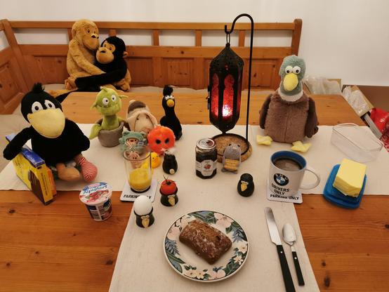 Ein gedeckter Frühstückstisch mit den beschriebenen Dingen, links sitzen ein Plüschrabe, der eine Packung blaugelbe Eszetschnitten hält, eine Monsterpflanze, ein Plüschschwein, ein Filzrabe, ein Plüschtentakel, ein Plüschfrosch, ein Plüschbärchen und zwei Gummienten. In Tischmitte steht eine rote, orientalische Kerzenleuchte, an deren Fuß eine hellgraue Häkelmaus  sitzt. Rechts der Lampe eine sehr große Plüschente. Munin (Rabenpfefferstreuer) zeigt uns beleidigt seine Rückseite. Ein Rabeneierbecher enthält eine Tomate, der andere ein Ei. Es gibt auch noch einen Rabensalzstreuer. Die Butterschale ist blau. Auf der Eckbank hinter dem Tisch sitzen zwei sich umarmende Plüschaffen.