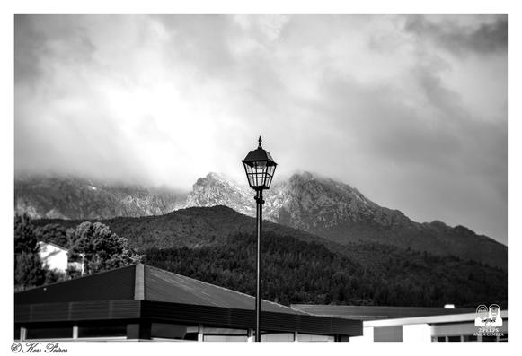 A moody black and white photograph featuring a classic street lamp centered against a dramatic mountain landscape, in Queenstown.

The foreground shows the roofline of a modern building. Behind the light, a forested slope rises to meet rugged, rocky peaks shrouded in low-hanging, textured clouds.

The overall composition creates a strong contrast between the man-made structure and the raw natural environment.