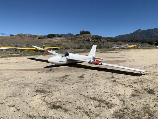 A Schweizer 1-26E glider, white with red and blue stripes, sitting on a dirt runway under clear, blue skies, a row of other gliders behind it.