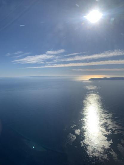 View over the Pacific Ocean towards the sun. The sea is calm, there are a few clouds. Santa Catalina Island is visible to the right, an in the far distance, San Clemente Island rises above the horizon