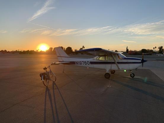 A bicycle (with fenders and pannier bags) and an airplane (a Cessna 172, white with blue and red stripes, stand on an airport apron at sunset