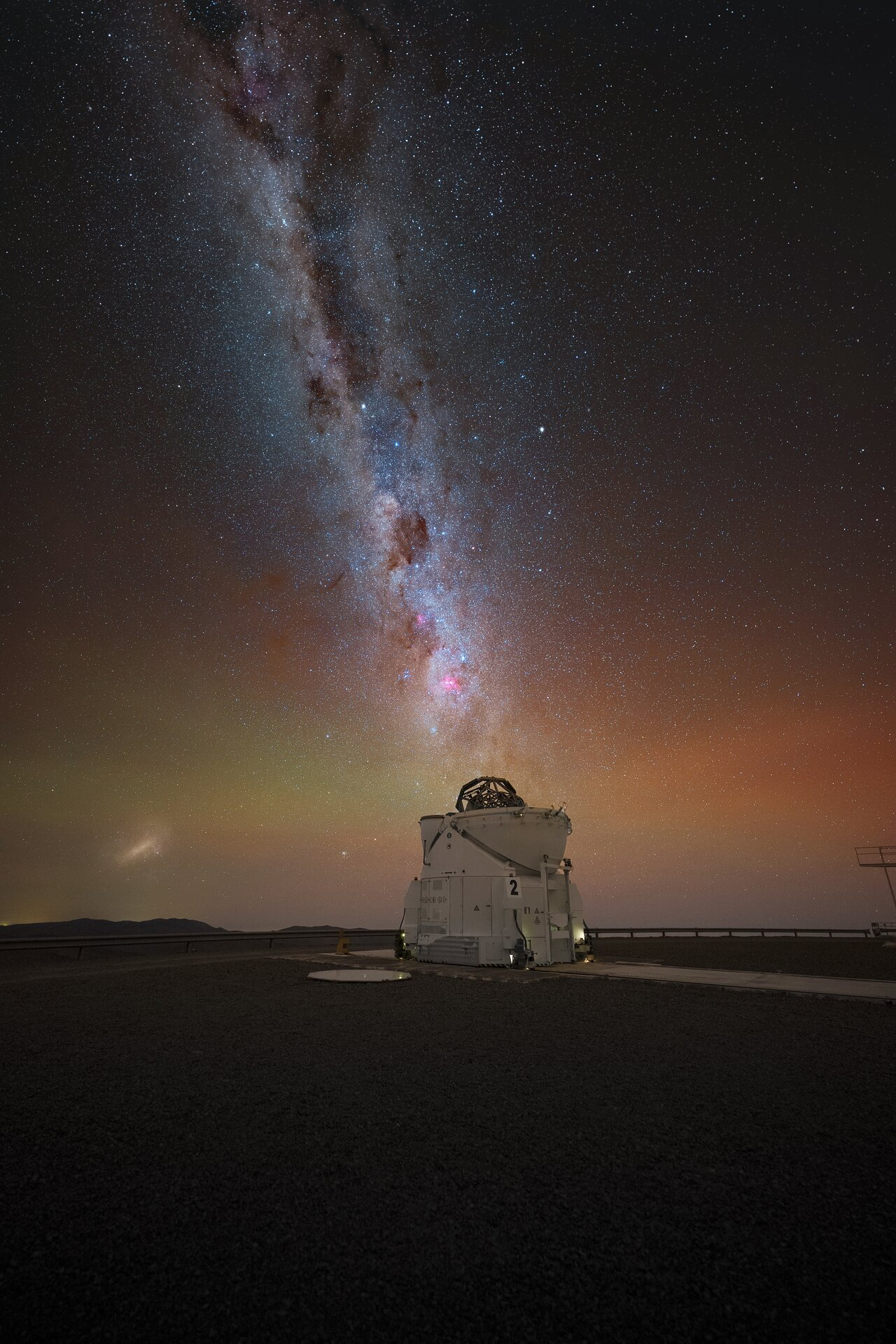 The image shows the Milky Way band seemingly pouring into a telescope, right below the centre of the image. Within the Milky Way right above the telescope, the Carina Nebula is visible as a pink spot, which marks the very centre and focus of the image. Additionally, one of the Magellanic Clouds is visible in the left side of the picture right above the horizon.