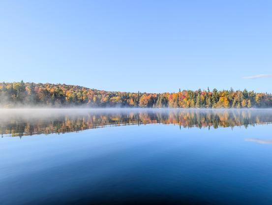 Mist rises over a tranquil lake, showcasing colorful fall foliage in the background.