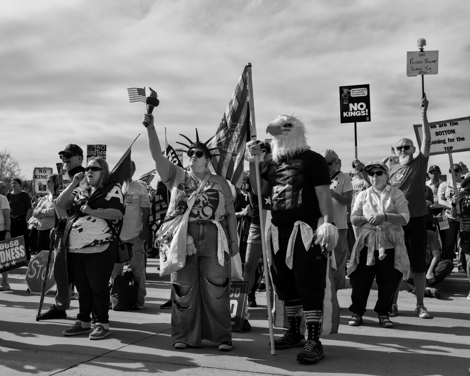 Black and white image of the No Kings Rally at the Iowa Capitol in Des Moines, Iowa. A person dressed as the Statue of Liberty stands next to a person dressed as Captain America with an eagle mask holding a US flag. Signs reading “No Kings!”, “We are the bottom coming for the top” and “Flush Trump Clean the bowl” can be seen in the crowd.