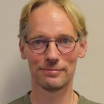 Foto van Frank, met glimlach, blonde haren met een scheiding. bril en baardje, kaki-groen shirt. 