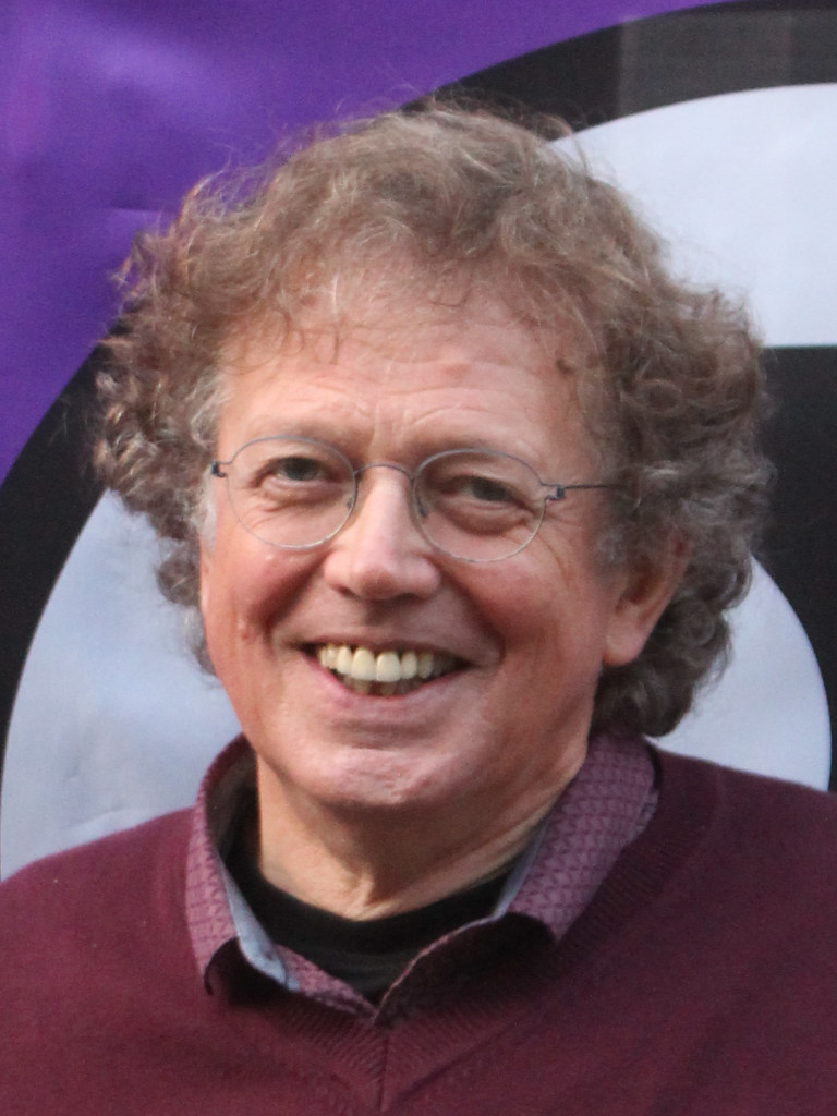 Foto van een lachende Edyy, bruin/grijs krullend haar, bril, rood-bruine trui met paarsig overhemd. Op de achtergrond is een deel van de Piratenvlag zichtbaar.