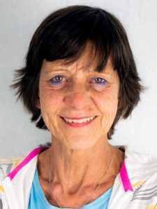 Op de foto een lachende Angeline met donker, kort haar, een wit jasje met roze streep en lichtblauw shirt. 