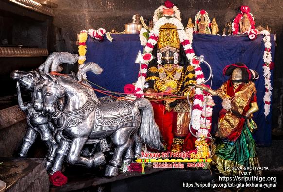 Idol of Hindu deity Krishna in red appears seated on a chariot driven by three horses that are all on Krishna's right; a silver arrow standing to Krishna's right with pointed end up; wearing gold shoes, gold palm coverings, and a gold crown. A string of white and red flowers around Krishna waist-waist. A woman ("satyabhaamaa") in red and green stands on the floor to Krishna's left and she holds out a silver bow. Dark blue cloth background. A string of white flowers hangs down at the right edge of the background. A string of yellow flowers hangs down at the left edge. 