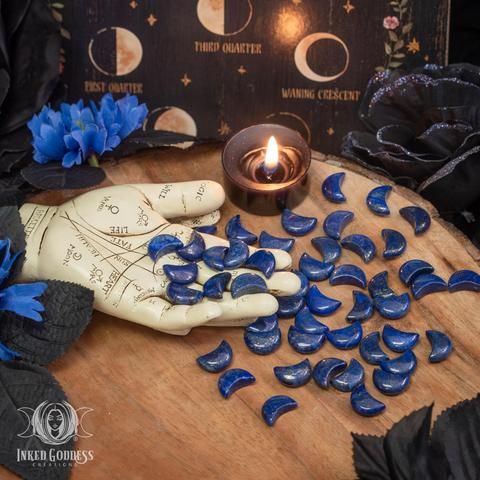 Many specimens of the "Lapis Lazuli Mini Crescent Moon" product are spilling from a palmistry figure. The moons are in a thick crescent shape and a deep, vibrant blue in color with pyrite inclusions. Their surface is polished smooth. Presented by Inked Goddess Creations.