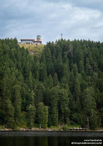 Perched atop a densely forested hillside, a quaint observation tower and building overlook a tranquil scene. The lush, evergreen forest—rich with deep greens and hints of lighter foliage—cascades down toward a calm, reflective lake below. The sky above is overcast, casting a soft, diffused light that mutes the colors into soothing tones, creating an atmosphere of quiet serenity.

The structure at the summit, partially obscured by the treetops, suggests a rewarding destination for hikers seeking…