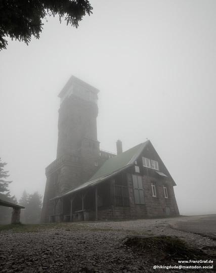Shrouded in a veil of soft, misty gray, a solitary stone tower rises from the foggy highlands, its rugged silhouette blending seamlessly with the muted tones of the overcast sky. The tower, sturdy and timeless, is accompanied by a quaint building with a green-tinged roof, its wooden bench inviting weary hikers to rest. The gravel path leading to the structure hints at countless journeys taken, each step a story of adventure and exploration.

The atmosphere is one of quiet mystery and contemplat…