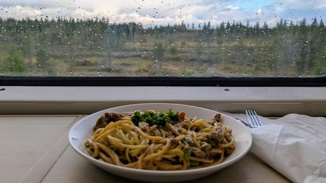 A plate with pasta on a lateral small table in front of a window, blurred heathland with trees outside, seen behind waterdrops
