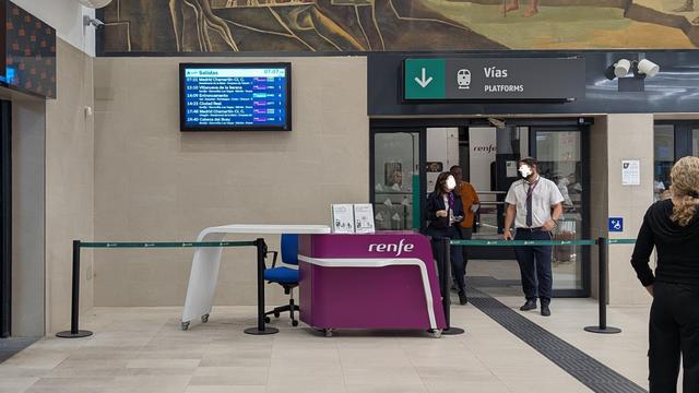 A waiting hall in a Spanish station, a Renfe desk with two Renfe staff members (anonymised) talking to each other behind the fenced off access to the platforms