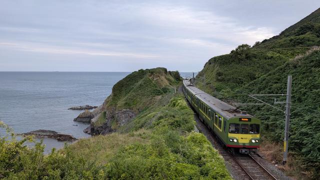 Two oncoming coupled DART 8100 class EMU on a bended track along a rocky and green coast line at dawn, some rocks in the sea