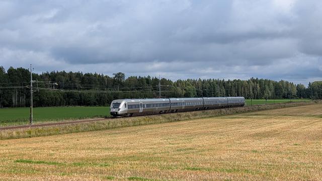 A SJ X55 Regina class EMU on a straight track, a harvested but still yellow appearing cereal field in front, a green meadow and a forest in the background, dark-grey clouds, sun-illuminated front of the train and foreground