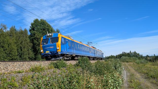 A Norrtåg X11 class EMU on a slightly bended track, a forest in the background, a trail parallel to the tracks, blue sky