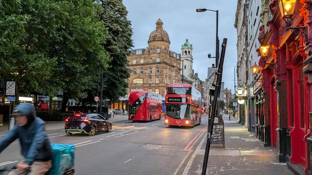 Oncoming red double decker bus not in service, red double decker bus in the opposite direction as line 19 at dusk, oncoming bike in front, historical buildings with cupolas in the background (edited image: number plates removed)