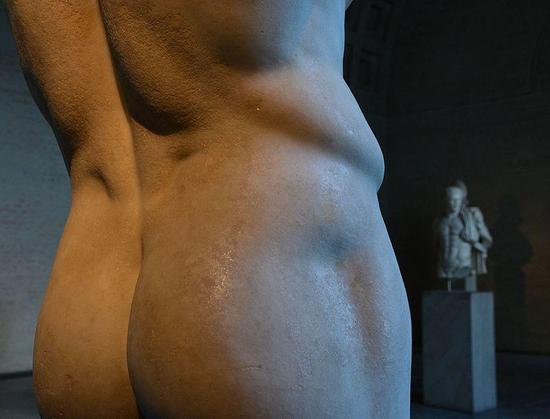 Detail of a sculpture's buttocks and lower back. In the background a marble torso of Herakles is averting his eyes.