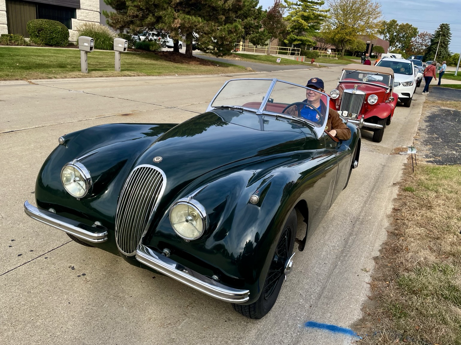 Photograph of a swoopy dark green Jaguar XK120 from the front. The car is parked on the side of a street with a bright red MG TC behind it.
