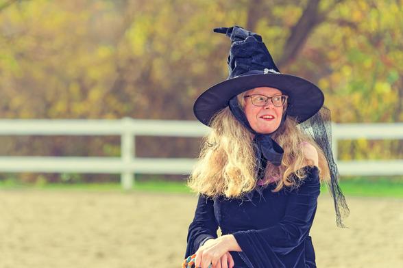 Blonde woman with long voluminous hair wears a witch hat tied on with a bow with a piece of chiffon hanging to the right with her arms folded with a white fence and fall color hedgerow in background