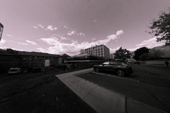 Black and white photo showing a distant and small looking concrete building in the center with a brick building that looks correspondingly sprawling towards the left and a parking lot to the right with white clouds clustered near the center of the horizon
