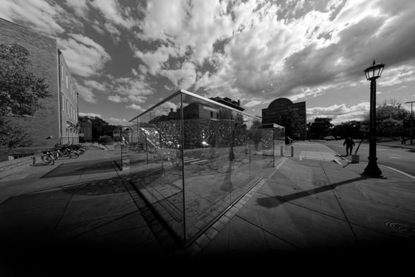The closest angle of the glass cube in front of us looks a lot sharper than 90 degrees because of wide angle distorsion,  confused with reflections of the environment can be seen a spreading stainless steel foam like the branches of a tree while a building looms to the left and a streelamp and road to the right under an unsettled sky