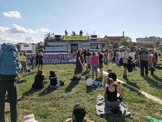 Eine Band spielt auf einem Reisebus auf einer großen Wiese, Menschen sitzen und stehen in kleinen Gruppen davor.
