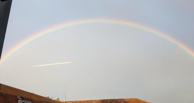 Ein Regenbogen, der direkt über einem Gebäude zu verlaufen scheint.

Unten im Bild rote Dachziegeln eines U-förmigen Gebäudes. Der Himmel ist grau bis zart blau.