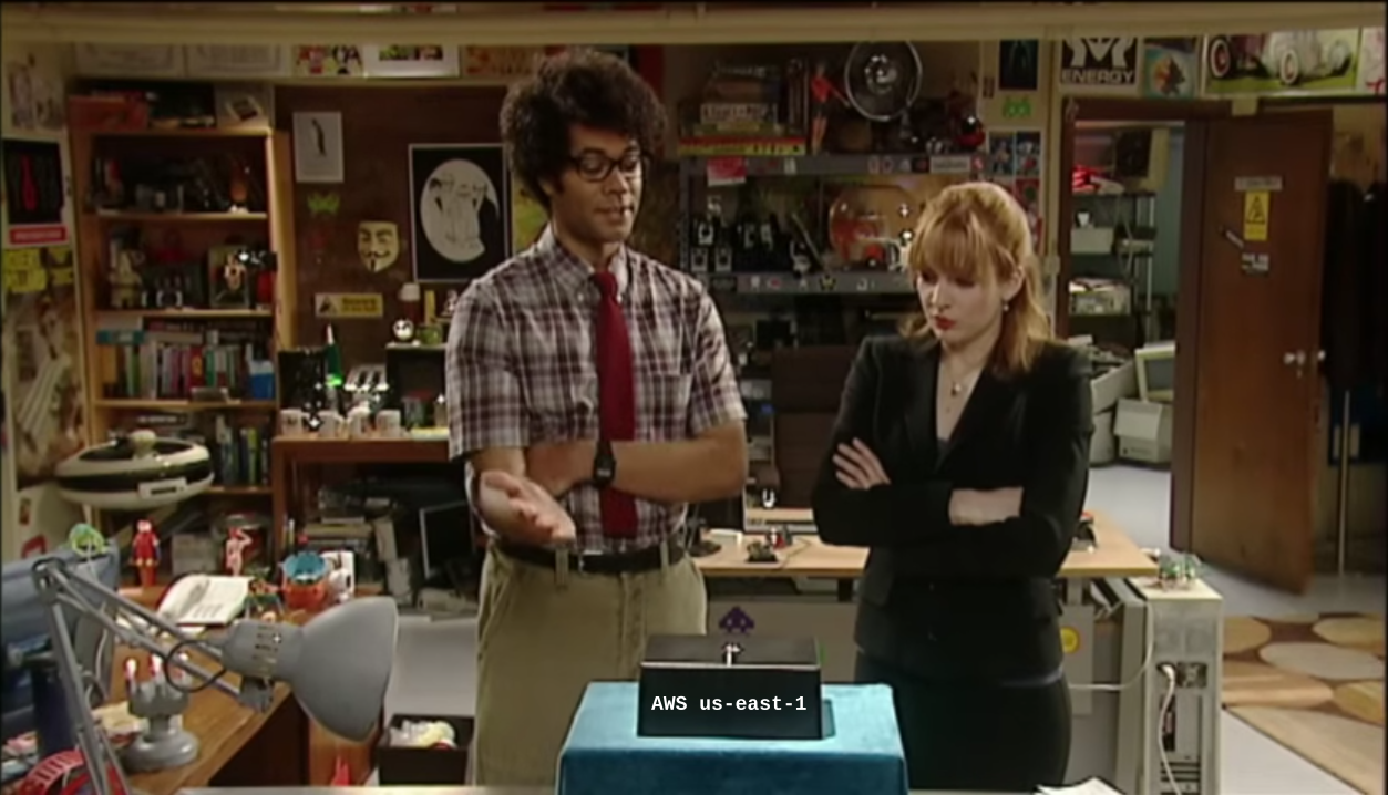 A scene from The IT Crowd where Moss tells Jen that a small black box on the table in front of them is the Internet, with the black box labelled AWS us-east-1.