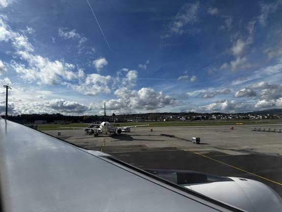 Blick über die Tragfläche, man sieht ein Triebwerk vorne herausschauen. Im Hintergrund parkierte Flugzeuge und Auto. Der Himmel ist stahlblau mit vielen weissen Wattebäuschen.