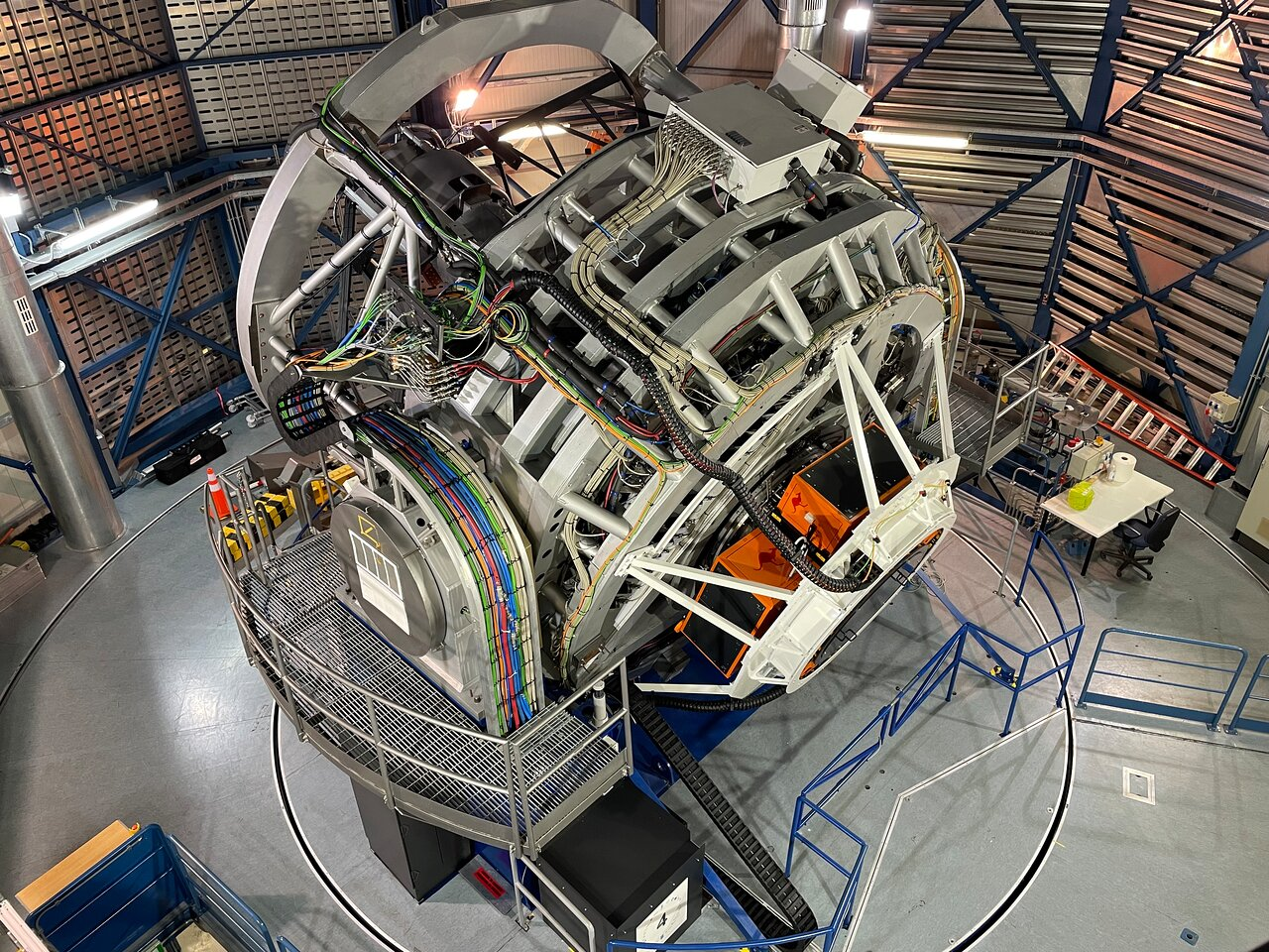 A view of the VISTA telescope and its 4MOST instrument. The structure is grey, with several cables visible around it. The telescope is viewed from above and is located inside a building with metallic walls.
