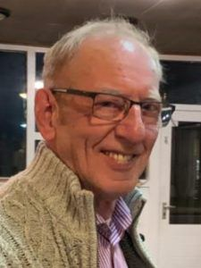 Foto van een lachende Teunis, kalend, lichtkleurig haar, bril, beige vest en wit-paars gestreept overhemd.