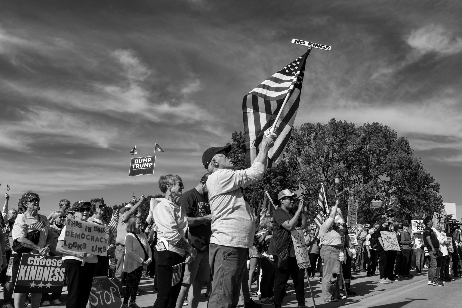 Black and white image of protesters at a No Kings Rally in Des Moines, Iowa. One man holds an American flag with a No Kings sign at the top of the pole.