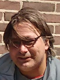 Foto van een lachende Peter, met beetje lang, sluik donker haar, bril en lichtblauw jasje