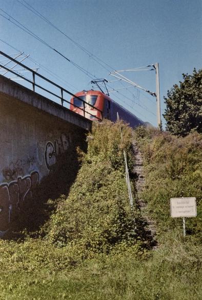 Viewed from below, a red locomotive drives on an elevated train track.