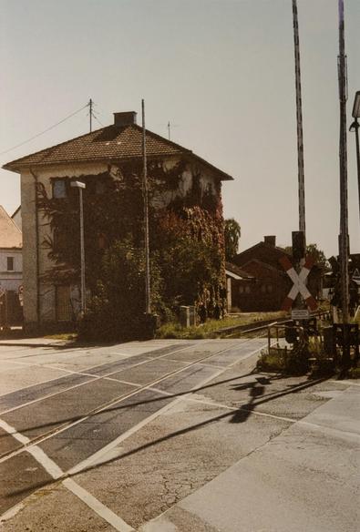 Behind train tracks of a railway crossing, a square building with a pointed roof is overgrown by colorful ivy in autumn