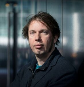 Foto van Dylan, sluik, donker haar, ringbaardje en donkerkleurig jasje op een wazige achtergrond.