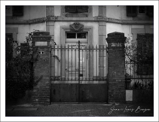 This black-and-white photograph captures a sense of age and quietude through its portrayal of an iron gate flanked by two sturdy brick pillars. The gate, slightly weathered but tall and imposing, bars entry to what appears to be an older building. The metalwork is simple, yet solid, with vertical bars topped by decorative points, hinting at a blend of security and subtle elegance.
Behind the gate, the building’s facade tells a story of a bygone era—the stone or plaster walls are framed with worn details and a small pediment above the door that evokes classical architectural influences. The doors themselves are stately, and their symmetry adds to the formal, restrained aesthetic. Framing the scene, the partially visible windows with closed shutters contribute to the sense of stillness and privacy.
The monochrome palette enhances the feeling of nostalgia and timelessness, stripping the scene down to contrasts and textures. The shadows play across the ironwork and brick, emphasizing the rugged solidity of the materials. The overall composition, with its balance between light and dark and its focus on the closed entrance, might evoke themes of secrecy, barrier, or a guarded past.
Artistically, this image could symbolize boundaries between the public and private spheres or the passage of time—the closed gate and aged building suggesting something precious or historical, quietly waiting beyond view.
