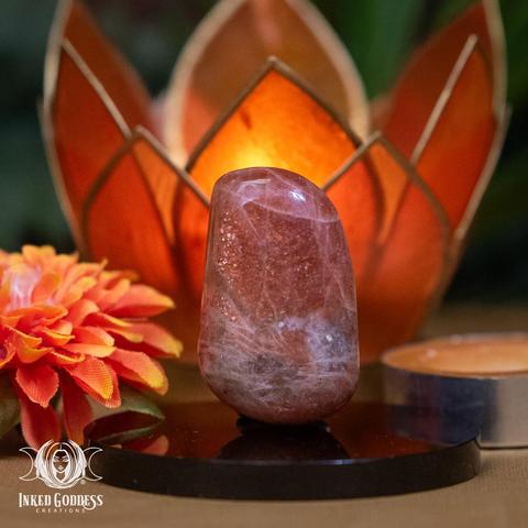 An example of the "Orange & Green Sunstone Tumbled Gemstone" product stands on an obsidian disc. This small tumbled gemstone is a glittery orange at the top and greenish at the bottom. It has irregular white veins. It's in a rough nugget shape and is polished smooth. Presented by Inked Goddess Creations.