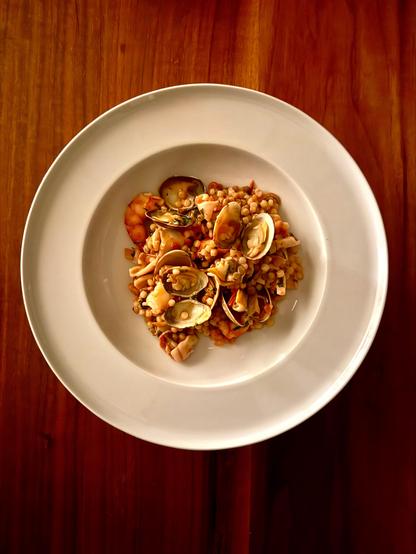 A deep white plate. Mussels, some with shells. Small squid and prawns. Fregola, a pearl-shaped pasta, covered with a reddish fish stock sauce.