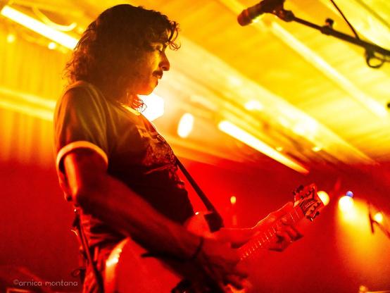 A musician plays an electric guitar on stage amidst dramatic lighting, featuring shades of orange and yellow. The image captures the performer in profile, highlighting their curly hair and focused expression.