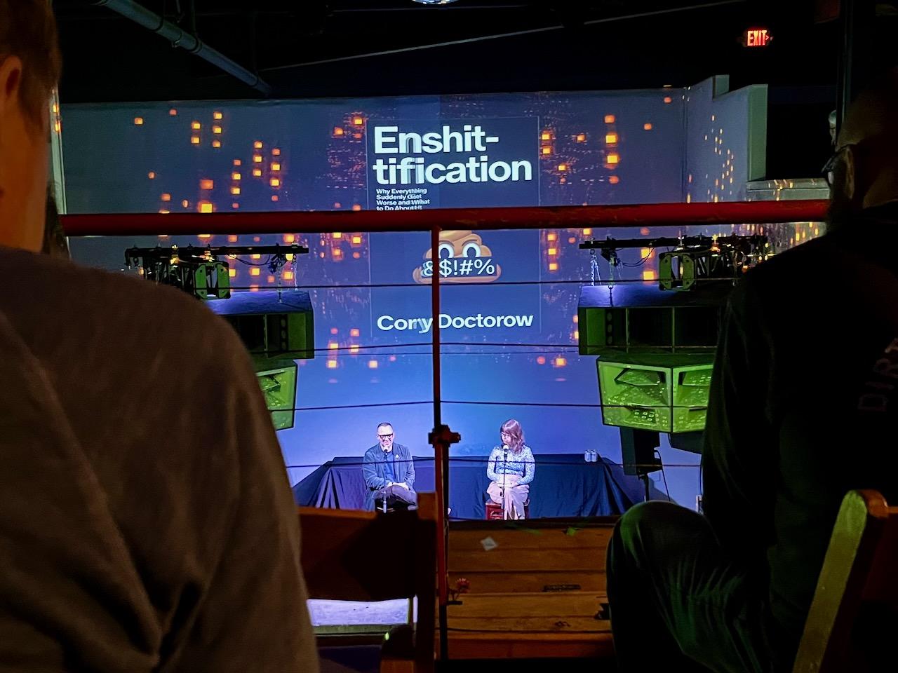 Cory Doctorow on stage with Jenny Odell. The title of Doctorow’s book, “Enshittification,” which features a an enraged “poop” emoji, is projected onto the wall behind the stage.