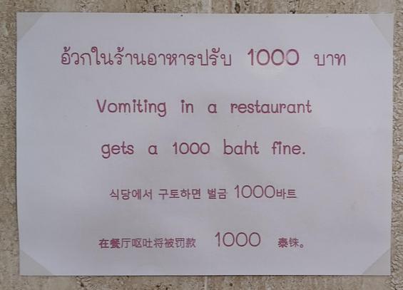 This is a photo of a simple, printed sign on a light beige textured wall, communicating a clear rule in multiple languages. The sign's main message, written in Thai, English, Korean, and Chinese, states that "Vomiting in a restaurant gets a 1000 baht fine." The text is printed in a maroon or dark pink color on white paper, which is taped to the wall.