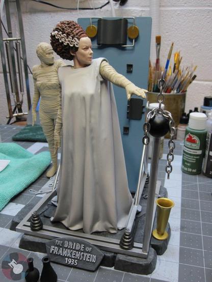 Model kit of the Bride Of Frankenstein by X-Plus. Gowned Bride in front of the procedure table, surrounded by metal framing and instruments. Standing on a stone floor with her left arm outstretched. Bandaged Bride in the background. On a workbench.