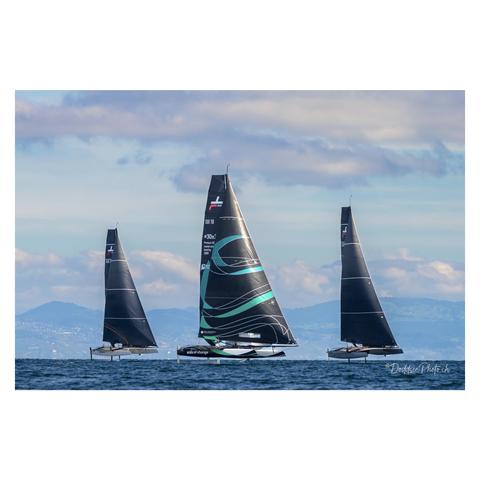 Three sleek black racing yachts sailing on calm blue water under a partly cloudy sky.