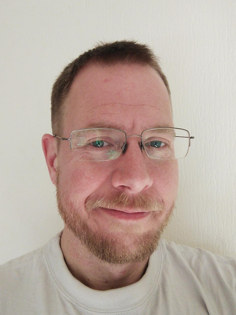 Foto van een lachende Dimitri, lichtrossig haar en snor, baardje, bril en wit shirt. 