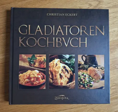 Foto des Gladiatoren-Kochbuchs auf Holzboden liegend - der Bucheinband ist braun, der Schriftzug "Gladiatoren-Kochbuch" in goldfarben, und darunter sind 3 Hochkant-Fotos von römischen Gerichten plaziert