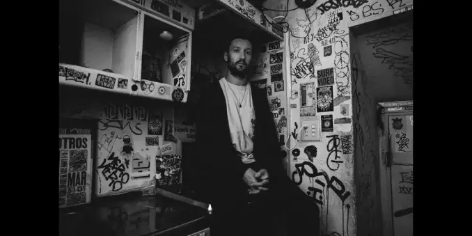 Bass player Max Luthert sitting in a graffiti-covered room, wearing a dark jacket and white shirt. Background adorned with stickers and posters, monochrome.